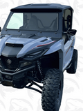 The Yamaha Rmax 1000 Glass Windshield is shown on a rugged, light blue off-road vehicle with large knobby tires, black grill, roof, angular headlights, high ground clearance, and a faint shark-patterned background.