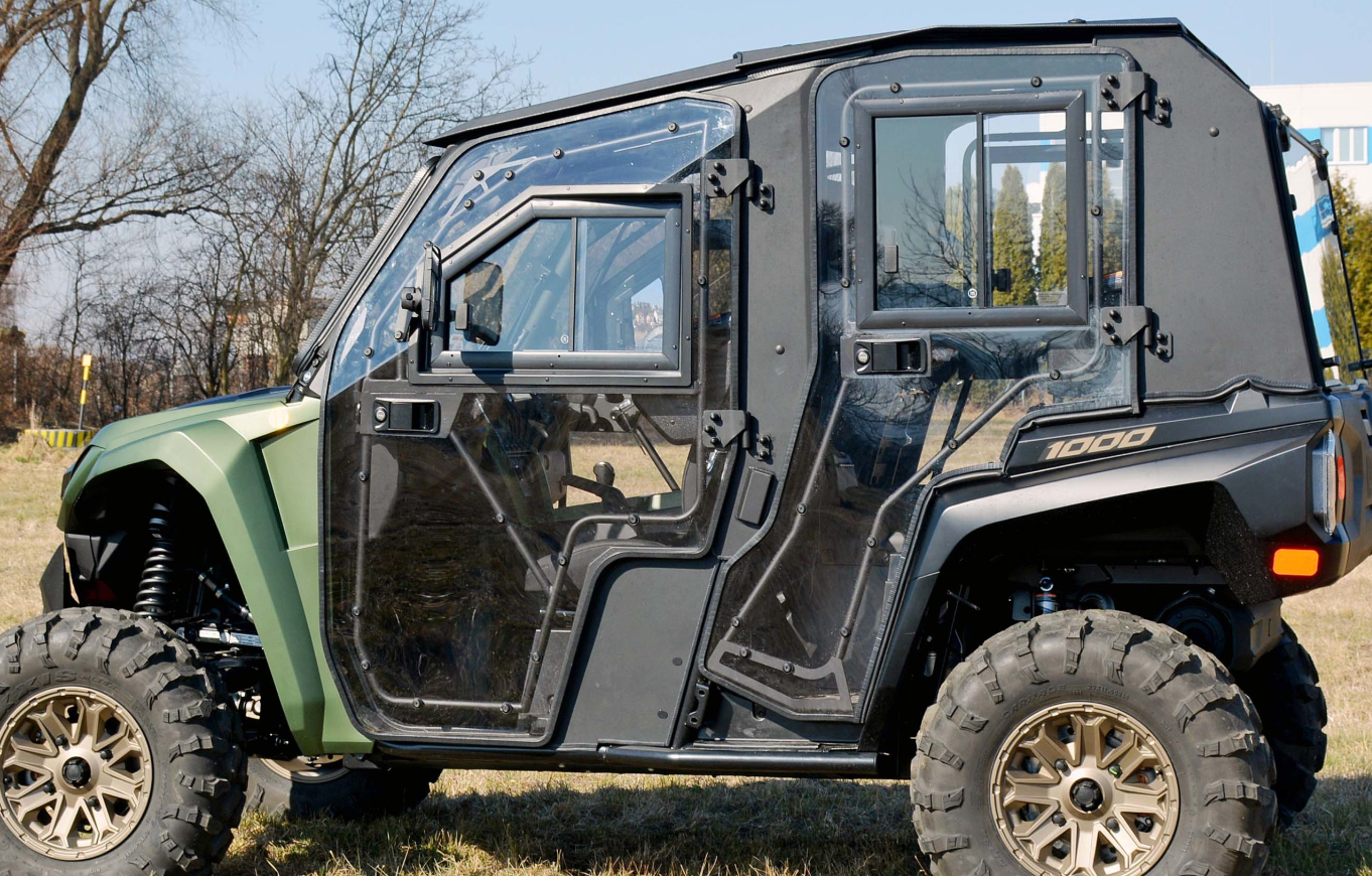A Yamaha RMax4 1000 Full Hard Cab Enclosure, black and green with large tires, is side-parked on grass. Its transparent angular doors, suspension, and 1000 mark are visible. Trees and a building are in the background.