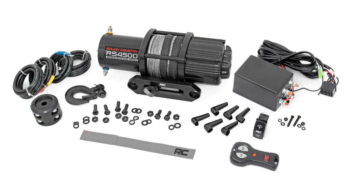 A complete Defender Game Loader kit is shown, featuring a black RC4WD RS4500 winch with cable, screws, nuts, bolts, hook, mounting brackets, wiring harnesses, controller box, remote control, and strap on a white background.