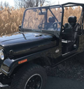 A black Mahindra Roxor with no doors and a roll cage, equipped with a Hard Coated Polycarbonate Windshield, is parked by tall dry grass; its two-seat interior is visible under a bright, sunny sky.