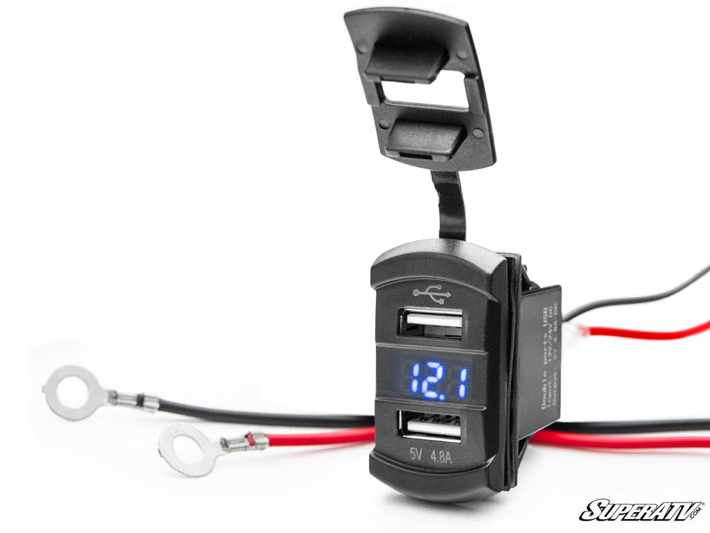 The 12 V UTV Voltmeter and Dual USB Charger displays 12.1 on its digital screen, features red and black wires with ring terminals, has an open dust cover, and shows the SuperATV logo in the bottom right corner.