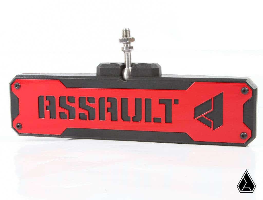 The Assault Industries Bomber Convex Center Mirror Bezel Set features a rectangular metal plate with a red front, black border, bold cut-out ASSAULT lettering, and a geometric logo on the right, attached to a mounting bolt.