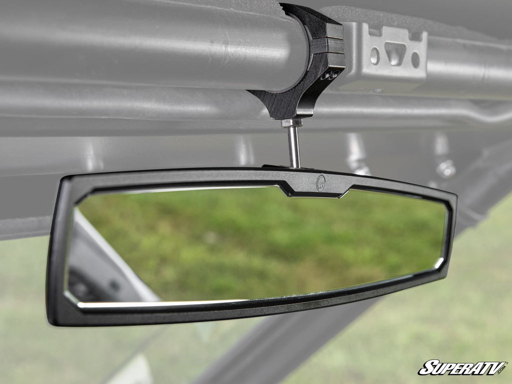 The Polaris RZR Aluminum Rear-View Mirror features a rectangular design with a black frame, mounted on a metal overhead bar. It reflects the vehicles interior and an out-of-focus green background, with the SuperATV logo in the lower right corner.