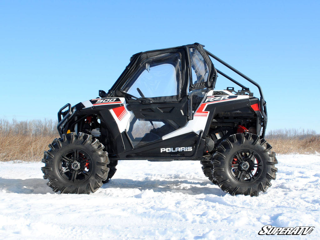 A Polaris RZR 900 equipped with the Polaris RZR 900 To RZR 900 S Conversion Kit With 3 Lift Kit, featuring rugged tires, red decals, and a clear enclosed cab, is parked on snow. The SuperATV logo appears in the bottom right corner.