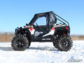 A Polaris RZR 900 equipped with the Polaris RZR 900 To RZR 900 S Conversion Kit With 3 Lift Kit, featuring rugged tires, red decals, and a clear enclosed cab, is parked on snow. The SuperATV logo appears in the bottom right corner.