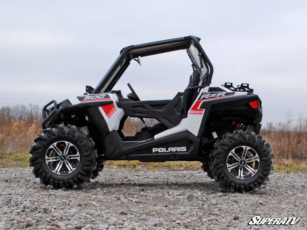 A Polaris RZR 900 equipped with the High Clearance 1.5 Offset Suspension Conversion Kit is parked on gravel. The open-cabin off-road vehicle, in black, white, and red, features large rugged tires with a cloudy sky and dry grass behind it.