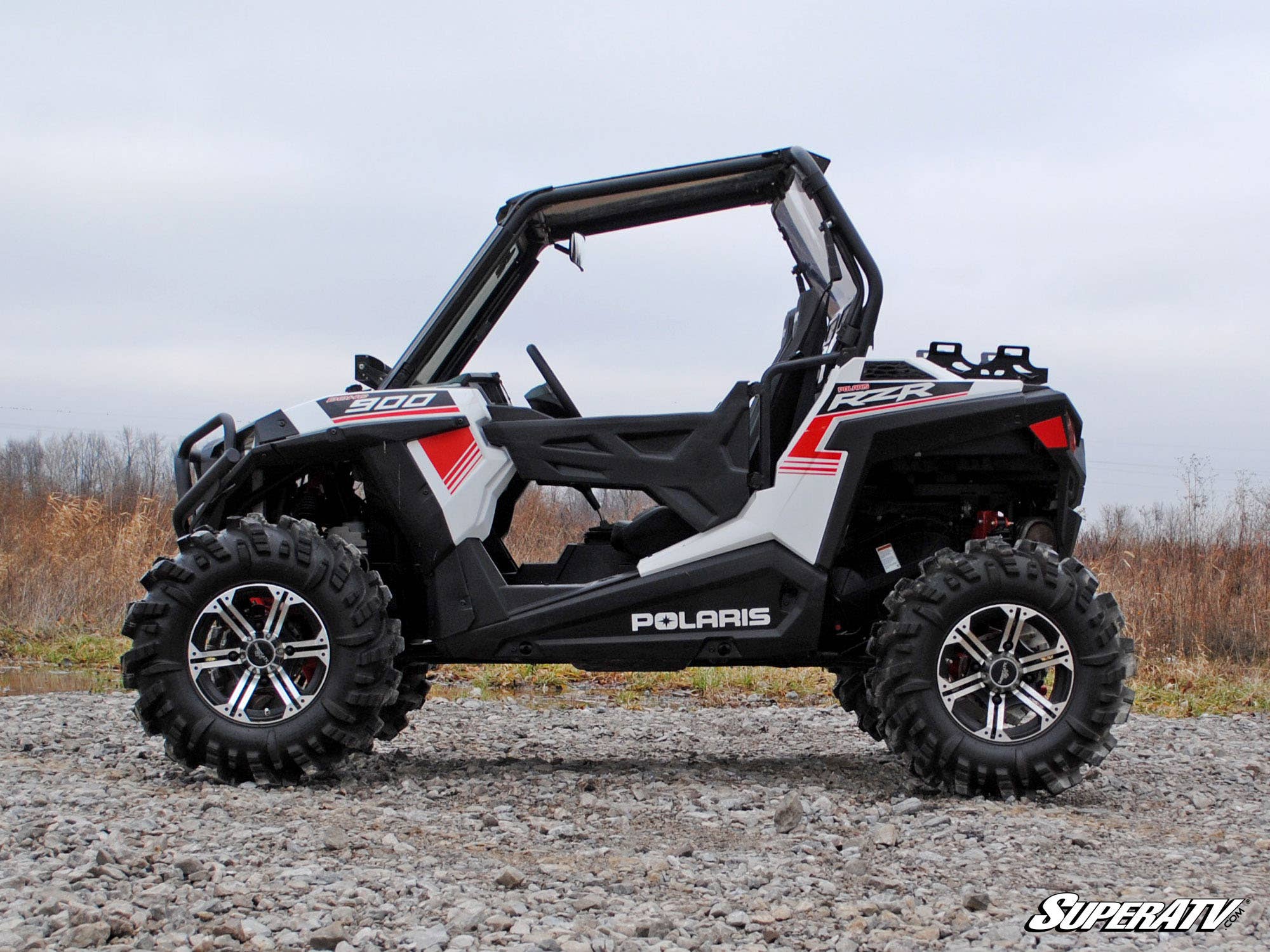 A Polaris RZR 900 equipped with the High Clearance 1.5 Offset Suspension Conversion Kit is parked on gravel. The open-cabin off-road vehicle, in black, white, and red, features large rugged tires with a cloudy sky and dry grass behind it.