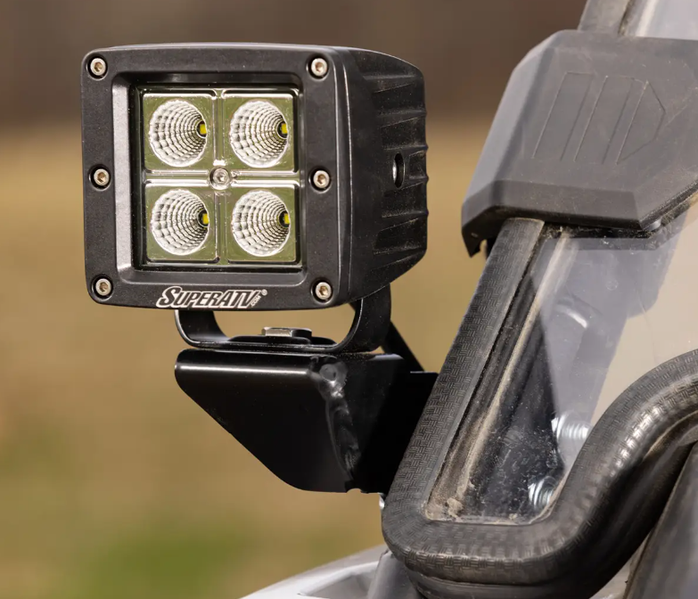 Close-up of Polaris RZR Cube Light Mounting Brackets holding a black LED light pod with four square-arranged lights, mounted beside a clear windshield with rubber edging on an off-road vehicle against a blurred natural background.
