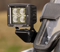 Close-up of Polaris RZR Cube Light Mounting Brackets holding a black LED light pod with four square-arranged lights, mounted beside a clear windshield with rubber edging on an off-road vehicle against a blurred natural background.