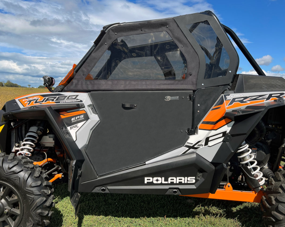 A Polaris RZR 1000XP with Solid Convertible Doors is parked on grass under a blue sky. The side shows Turbo, XP, and Polaris branding, a strong suspension system, and transparent convertible doors with black frames.