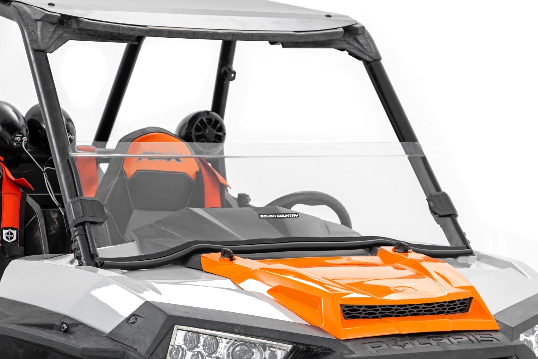 A front close-up of a Polaris RZR XP 1000 (2014-18) with a Rough Country Half Windshield (scratch-resistant); the clear windshield, visible roll cage, orange and black seat upholstery, headlight, and hood are all in view.
