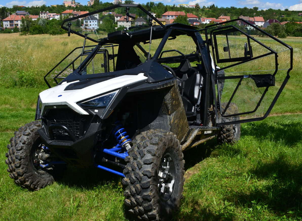 A Polaris RZR Pro XP Cab Kit, featuring large muddy tires and an angular design, is parked on grass with its doors open to show a black-and-white interior. A field and hilltop houses appear in the background under a blue sky.