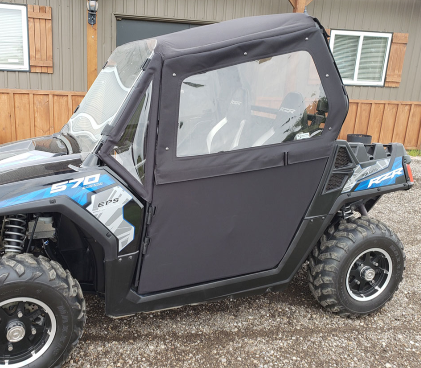 A side view of a black and blue RZR with the Polaris RZR Complete Cab Enclosure Kit With Full Windshield, including soft black doors and windows, parked on gravel in front of a wooden building with shuttered windows.