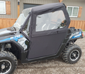A side view of a black and blue RZR with the Polaris RZR Complete Cab Enclosure Kit With Full Windshield, including soft black doors and windows, parked on gravel in front of a wooden building with shuttered windows.