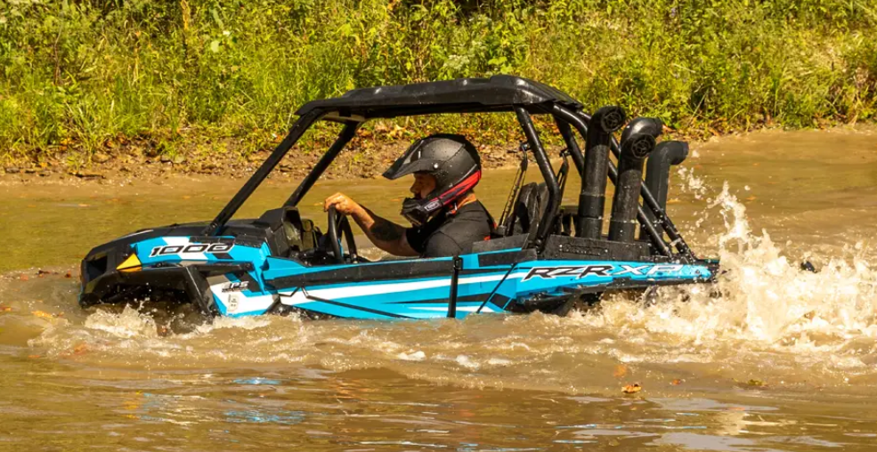 A helmeted rider drives a Polaris Rzr equipped with the Pro XP 1000 Depth Finder Snorkel Kit through a shallow, muddy creek, creating splashes. Lush greenery and tall grass line the banks in the background.