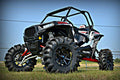 A Polaris Rzr Xp 1000 with the S3 Power Sports 5 Bracket Lift Kit, black and white with oversized mud tires, red suspension, and a roll cage, is parked on grass under a blue sky, viewed from a low angle.