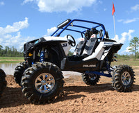 A Polaris Rzr Xp 1000 with an S3 Power Sports 2014+ 8 Lift Kit features large mud-terrain tires and chrome wheels, parked on dirt by shallow water under a bright sky. An orange flag is attached to the roll cage in the background.