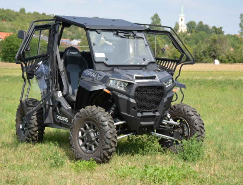 A Polaris RZR XP1000 Turbo (2019) Hard Cab Enclosure, featuring rugged tires, a protective frame, transparent doors, and a windshield, is parked on grass with trees, a field, and a church steeple in the background.