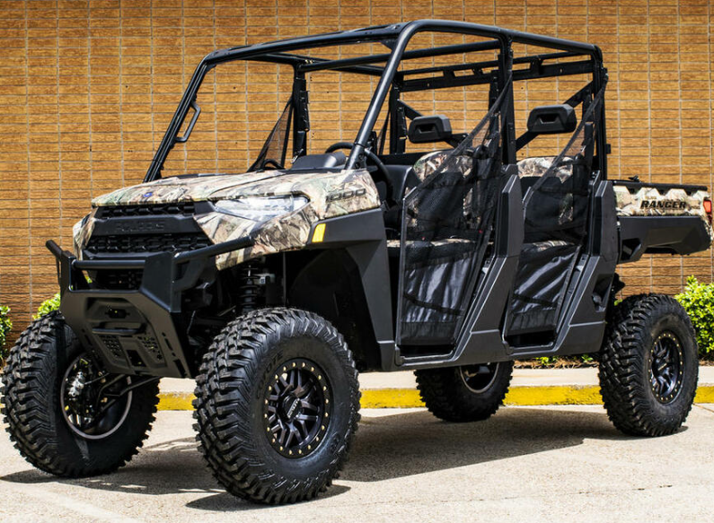 A Polaris Ranger equipped with the S3 Power Sports 2018-2020 4 Lift Kit, featuring a rugged camo pattern, off-road tires, four doors with mesh panels, and a cargo bed, parked before a tan brick wall and green shrubs.