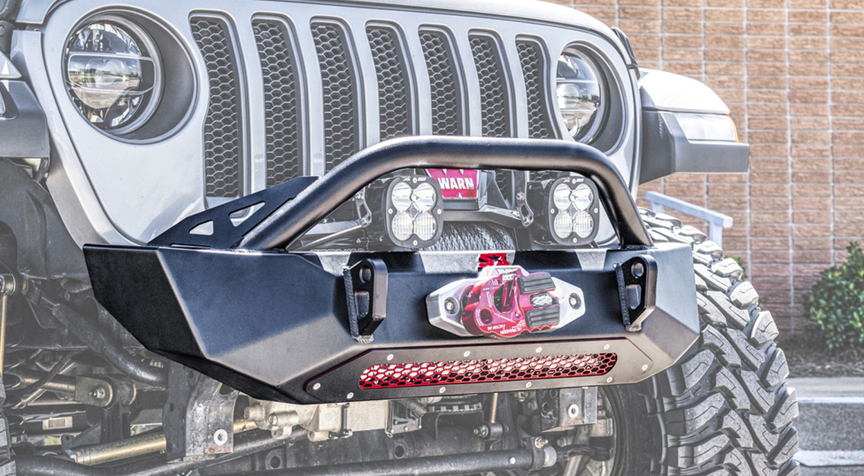 Close-up of a Jeep Wrangler JK/JL/JT Ambush Series Front Winch Bumper in black, featuring an integrated winch, red tow hook, off-road lights, metal grille, and large tires. Brick wall and small plants appear in the background.