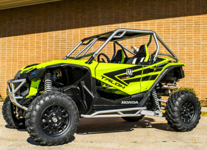 A bright green Honda Talon with S3 Power Sports 2019+ Honda Talon Nerf Bars, black accents, roll cage, large rugged tires, and roof bars is parked on a driveway in front of a tan brick wall. Two bucket seats are visible inside.