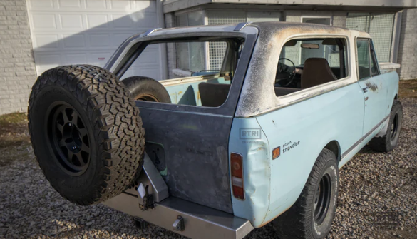 A light blue vintage International Scout with a weathered exterior and open rear cargo door reveals the inside-mounted spare tire. Parked on gravel by a white garage, its worn roof and paint show rust—ideal for showcasing our Rear Bumper for International