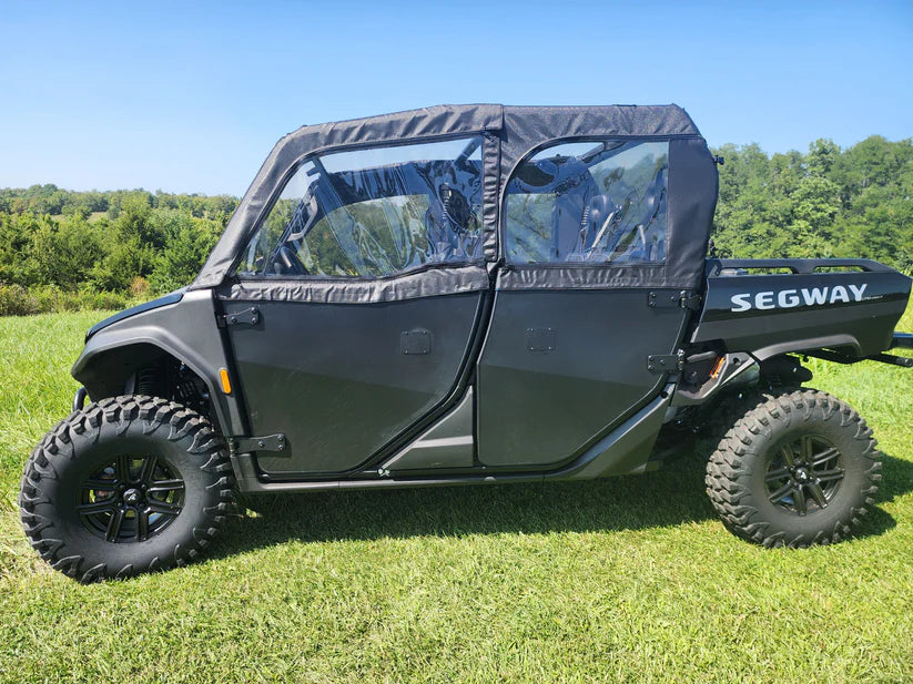 A Segway UT10 Crew with soft doors, large rugged tires, and clear windows is parked on green grass. The black off-road utility vehicle features two doors and a visible cargo bed, set against trees and a blue sky backdrop.