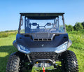 A Segway UT10 Crew with a 1-piece scratch-resistant front windshield featuring vents is parked on green grass, viewed head-on with rugged tires and LED headlights. Trees and a blue sky are in the background.
