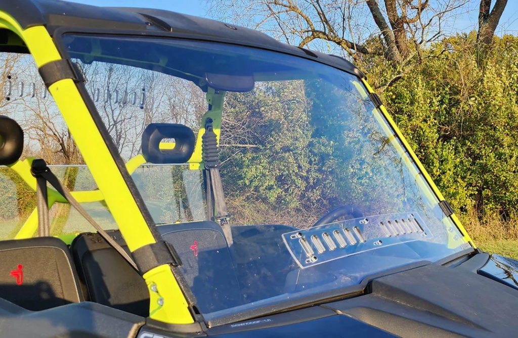A close-up of a Segway UT10 shows its scratch-resistant windshield, black seats with headrests, and sunlight reflecting on the glass. Green bushes, leafless trees, and a blue sky are visible behind the vehicle.