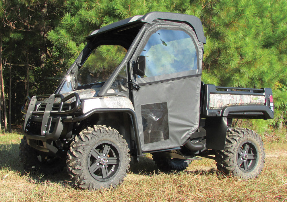A side view of a John Deere Gator Full Size XUV with a Hinged Door Kit, large rugged tires, roof, and enclosed sides parked on grass. Dense green pine trees are in the background. The vehicles bed is partially open, highlighting its utility.