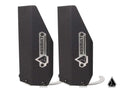 Two black, angular metal Assault Industries Extended Shock Guards // Front with silver logo plates and geometric icons stand upright side by side. Visible screws and curved hooks are at the bases. A smaller logo appears in the lower right corner.