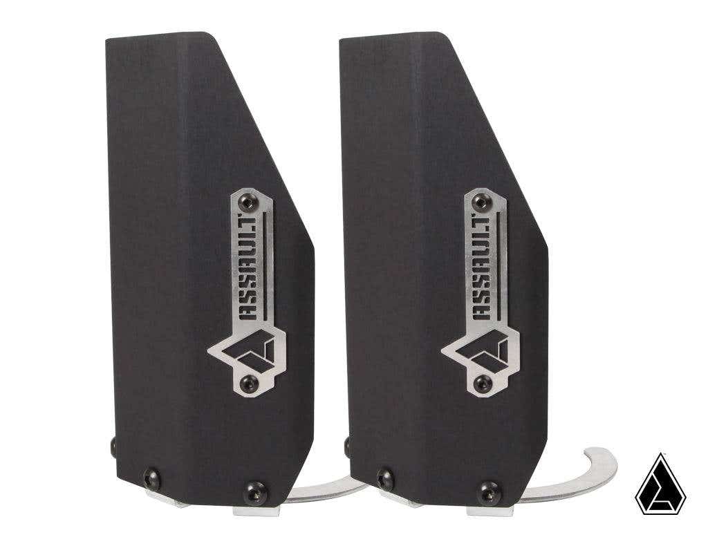 Two black, angular metal Assault Industries Extended Shock Guards // Front with silver logo plates and geometric icons stand upright side by side. Visible screws and curved hooks are at the bases. A smaller logo appears in the lower right corner.