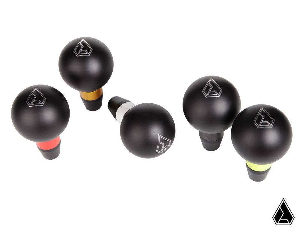 Five Assault Industries GT Shift Knobs for Can-Am Maverick X3, each with a silver geometric logo and a colored band (red, orange, white, green, yellow) at the base, are arranged on a white background. A matching triangle logo is in the bottom right corner