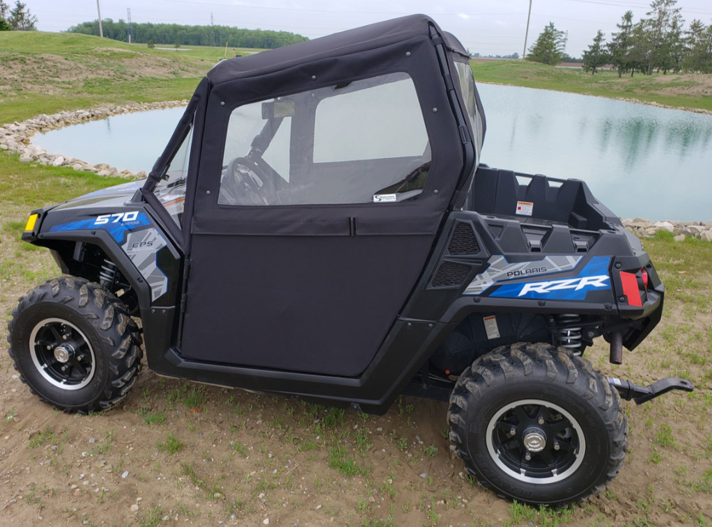 A black Polaris RZR with Convertible Doors for 570/800/900 models is parked on dirt near a pond, featuring large rugged tires and “570” and “RZR” decals. Trees, grass, and a stone border surround the water in the background.