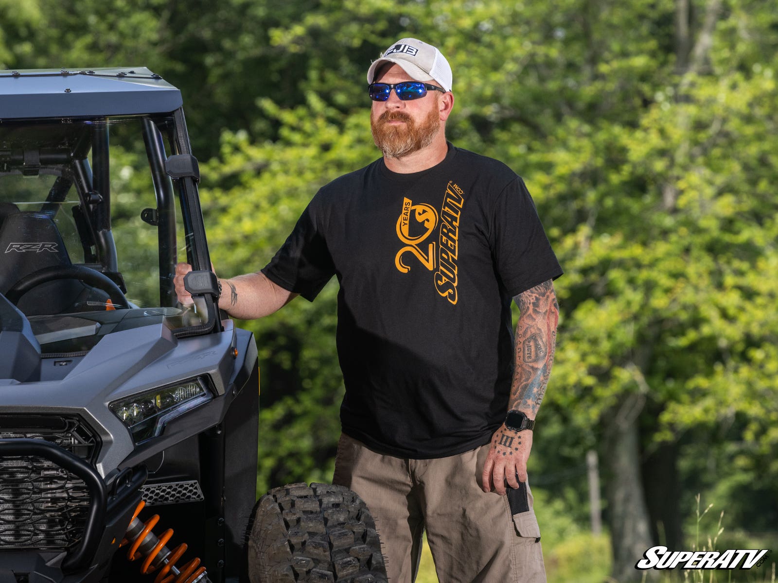 A bearded man in sunglasses, a white cap, a smartwatch, and an Orange & Black 20th Anniversary T-Shirt stands outdoors by an off-road vehicle with trees in the background. The SuperATV logo appears in the bottom right corner.