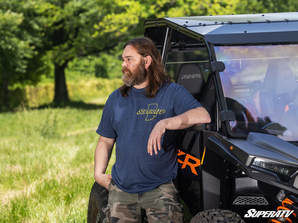 A man with long brown hair and a beard, wearing a Blue & Yellow 20th Anniversary T-Shirt and camouflage shorts, leans against a black off-road vehicle in a grassy field. The SuperATV logo appears in the bottom right corner.