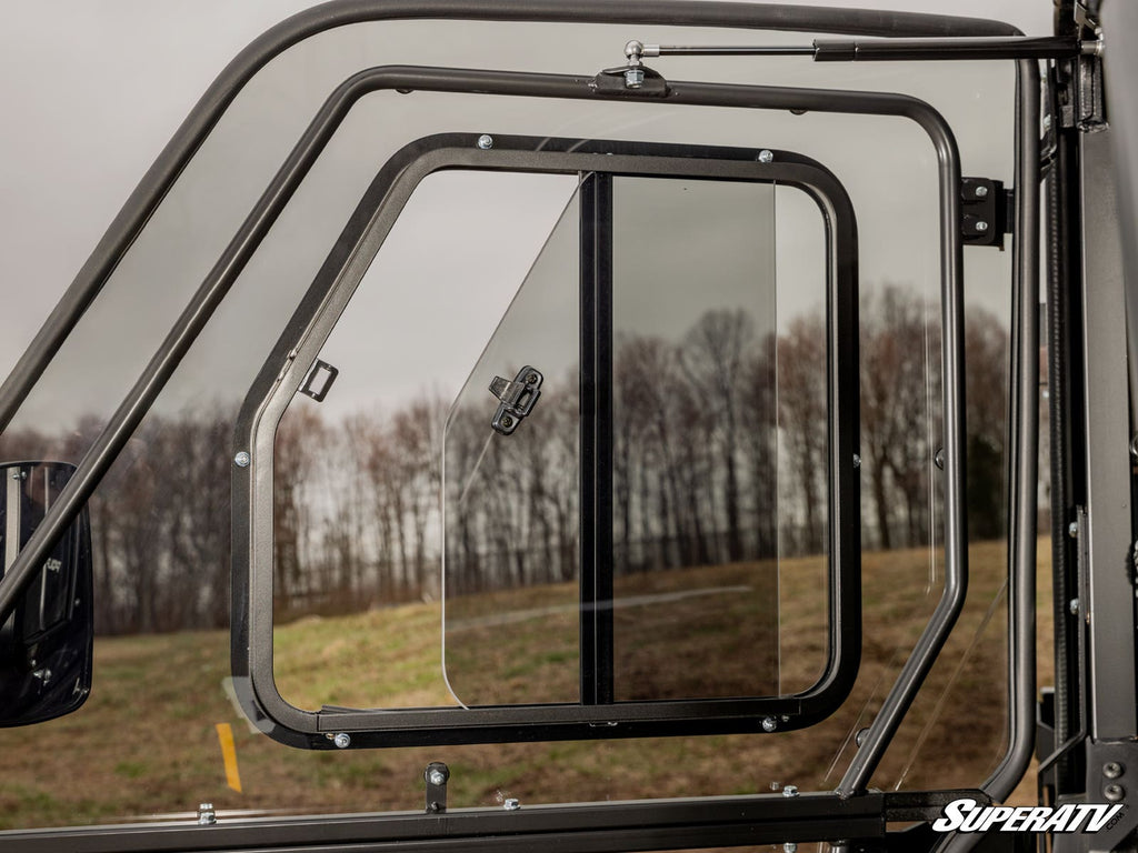 Close-up of a partially open Sliding Window Insert for SuperATV Poly Cab Doors, featuring a rectangular sliding panel in black metal framing. Blurred trees and grass appear outside under a cloudy sky, with the SuperATV logo at bottom right.