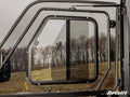 Close-up of a partially open Sliding Window Insert for SuperATV Poly Cab Doors, featuring a rectangular sliding panel in black metal framing. Blurred trees and grass appear outside under a cloudy sky, with the SuperATV logo at bottom right.