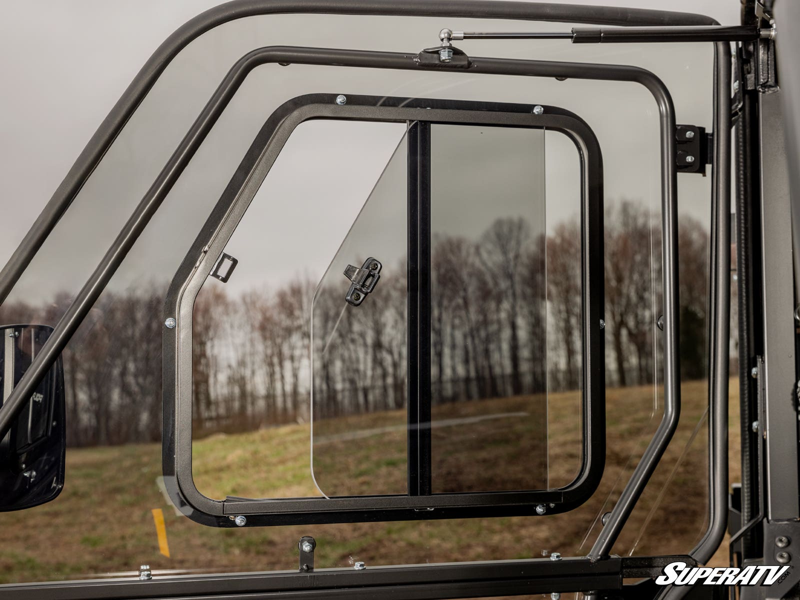 Close-up of a partially open Sliding Window Insert for SuperATV Poly Cab Doors, featuring a rectangular sliding panel in black metal framing. Blurred trees and grass appear outside under a cloudy sky, with the SuperATV logo at bottom right.