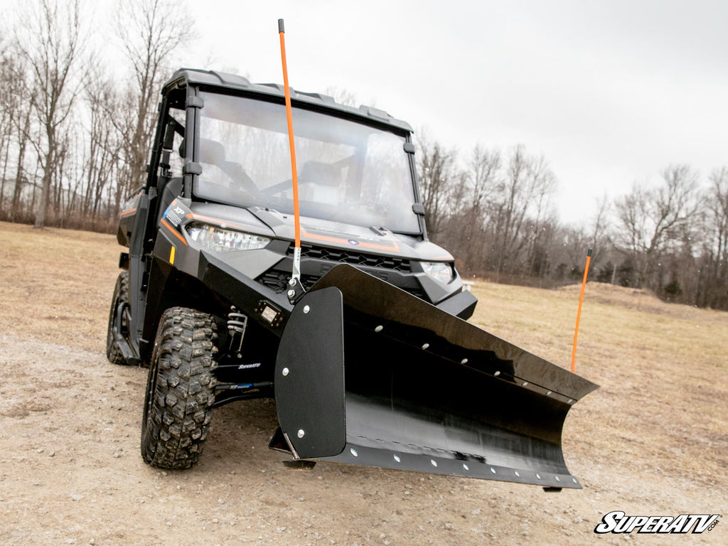 A black off-road utility vehicle equipped with the Plow Pro Snow Plow Deflector and Marker Kit is parked on a gravel path. Orange markers are mounted on the plow. Leafless trees and grass appear in the cloudy background.