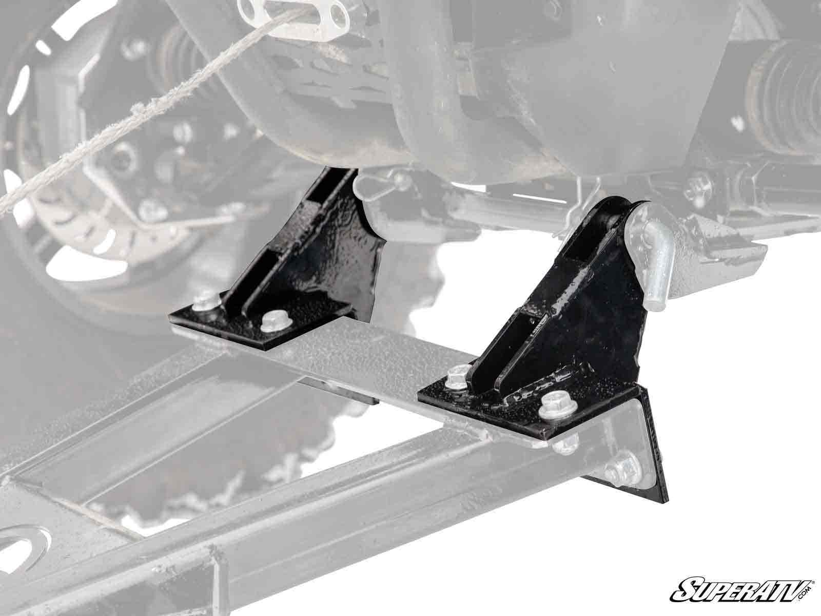 Close-up of sturdy, angular Plow Pro Snow Plow Drop Brackets in black metal, mounted to a vehicle frame with four bolts. The brackets are highlighted against a faded background; SuperATV logo appears in the bottom right corner.