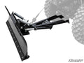 A close-up of the Plow Pro Snow Plow Blade and Frame Kit in black metal, attached to an off-road vehicle with rugged tires. The blade features tension springs, a mounting frame, and displays the SuperATV logo at the bottom right. Vehicle is partially visi
