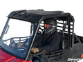 A person wearing a black helmet, goggles, and gloves drives a red off-road vehicle featuring the Polaris Ranger Mid Size 570 Plastic Roof (2015+) with netted doors. The scene has a plain white background and SuperATV branding in the bottom right corner.