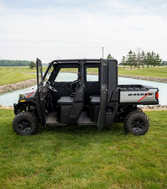 A Polaris Ranger 570SP Crew with Solid Convertible Doors, shown in black with open driver and rear passenger doors, is parked on grass beside a canal, with trees and a cloudy sky in the background.