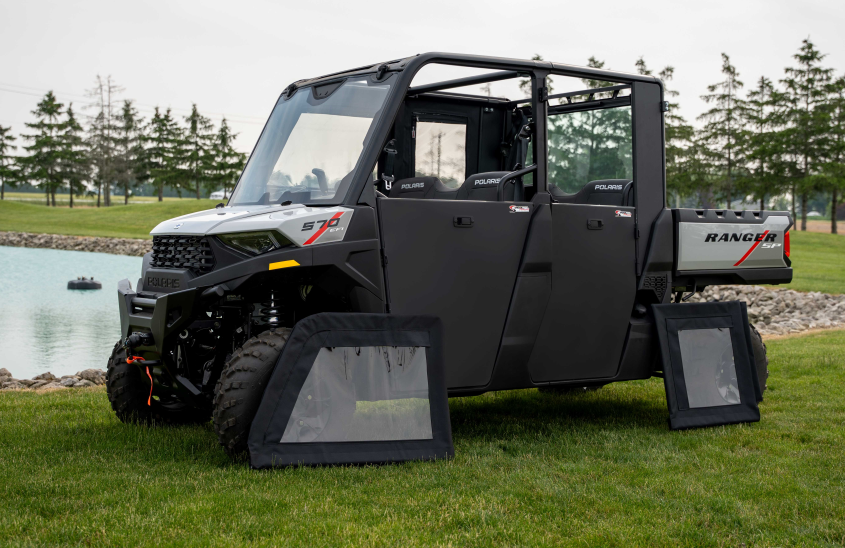 A Polaris Ranger 570SP with Crew Solid Convertible Doors is parked on grass by a pond. Two clear, detached doors lean against it, while the left front and rear doors are open. Pine trees and power lines appear in the background.