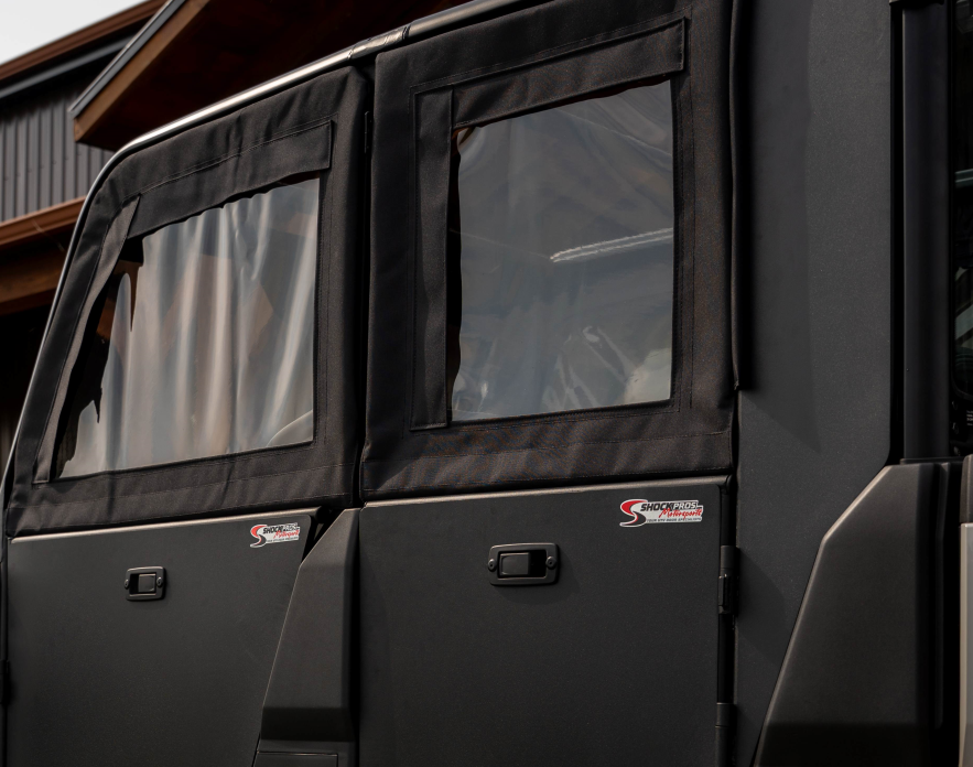 Close-up of Polaris Ranger 570SP Crew Solid Convertible Doors with clear plastic windows, rectangular metal handles, and red-and-white “Shockey Motorsports” logos. A wooden building is partially seen in the background.