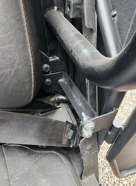 Close-up of a Polaris Ranger 570SP Crew Solid Convertible Door shows a seatbelt mounted to a metal frame beside a padded seat. Bolts and washers secure the bracket, with dusty surfaces and part of a barcode label visible. Gravel ground appears outside.