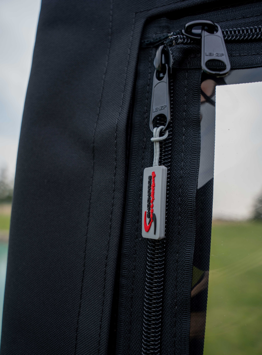 Close-up of a black zipper on Polaris Ranger 570SP Crew Solid Convertible Doors, with a rectangular pull showing the red and black Switchback logo. The blurred background displays green grass and faint tree outlines.