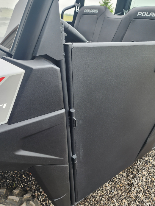 Close-up of a Polaris Ranger 570SP Crew Solid Convertible Door in matte black, slightly open to reveal two POLARIS-labeled seats. The utility vehicle is parked on small gravel stones.