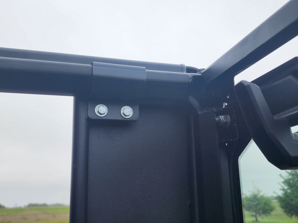 Close-up of a Polaris Ranger 570SP Crew Solid Convertible Door’s metal frame corner, showing two bolts on a black hinge bracket. Background: cloudy sky, grassy field, distant trees, visible through the partly open door window.